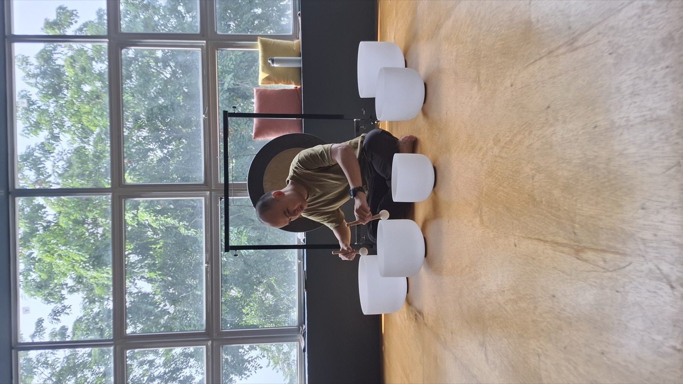 Hong seated behind a row of crystal singing bowls with a gong, in a daylit studio